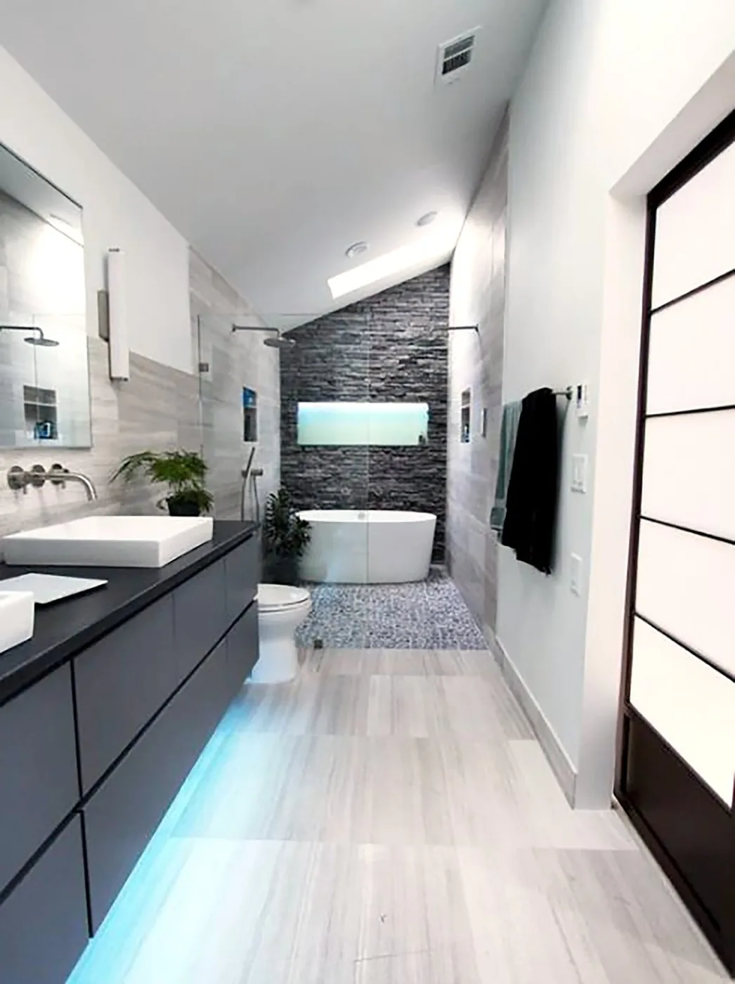 Matte black floating double vanity with integrated finger-pull edges and under-cabinet LED glow, topped by twin white vessel basins and wall-mounted chrome faucets against a stacked stone feature wall