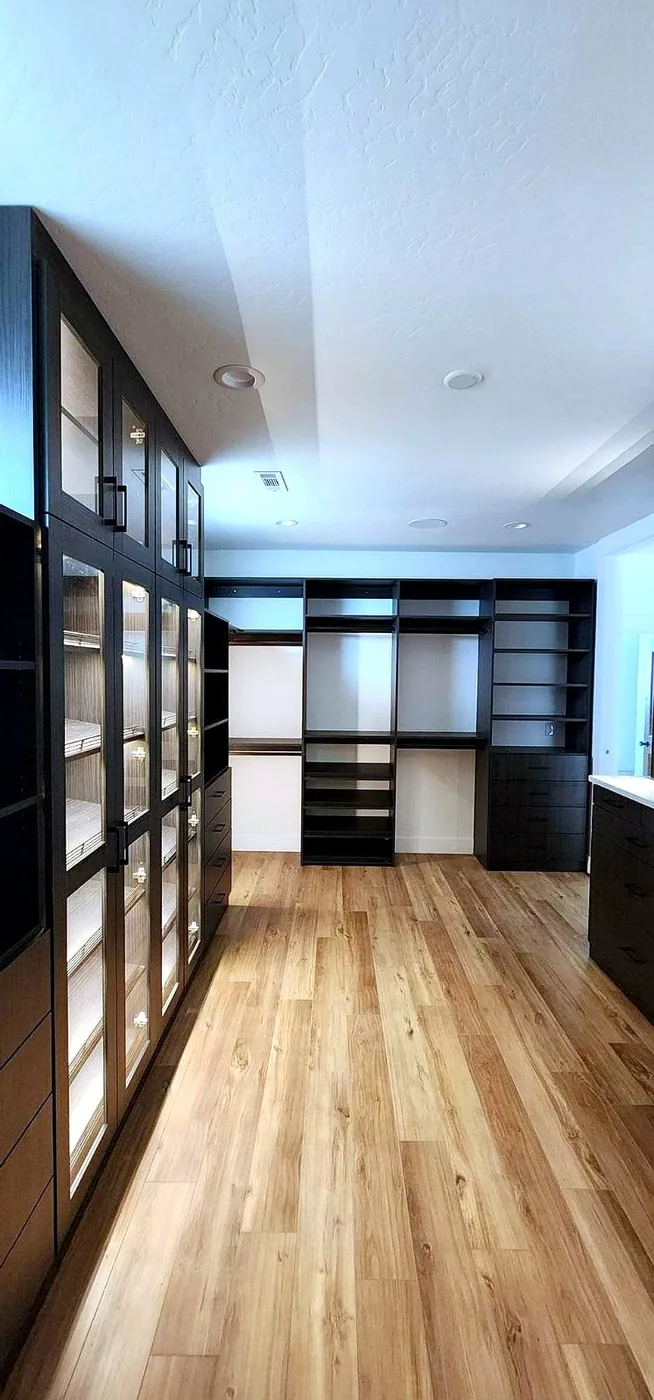 A dramatic espresso-finished walk-in closet lined with LED-lit glass display cabinets, open shelving and hanging rods running toward a bright window