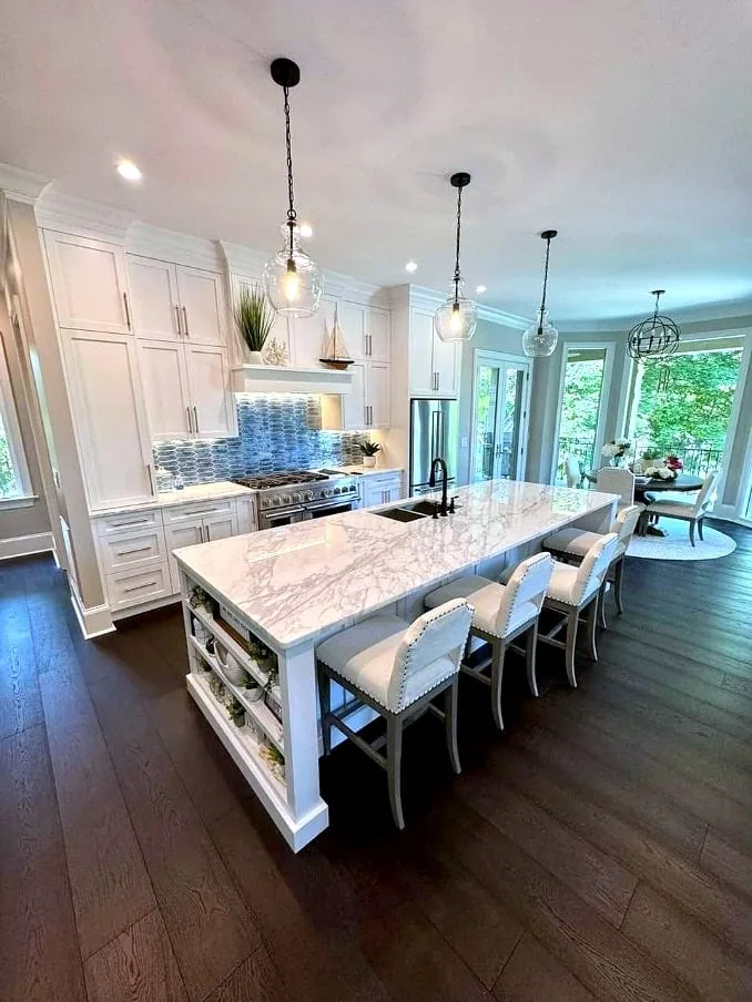 Full-height white cabinetry with a panelled range hood and blue mosaic backsplash surrounds a long marble waterfall island seated for five, with glass globe pendants above and dark hardwood underfoot