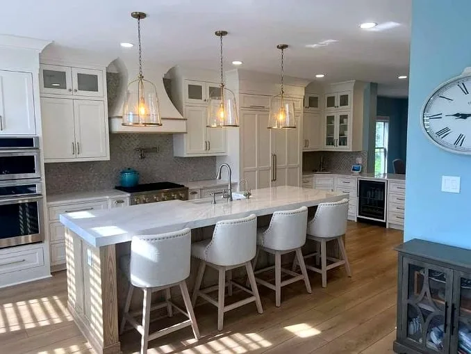 Cream inset cabinetry with a curved plaster range hood, mosaic backsplash and glass-front uppers wraps a marble-topped island seated for four, lit by three brass-and-glass lantern pendants over wide oak flooring