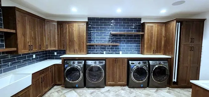 Warm walnut shaker laundry cabinetry with open floating shelves, a navy subway-tile backsplash, white farmhouse sink and two stacked stainless washer-dryer pairs on white counters