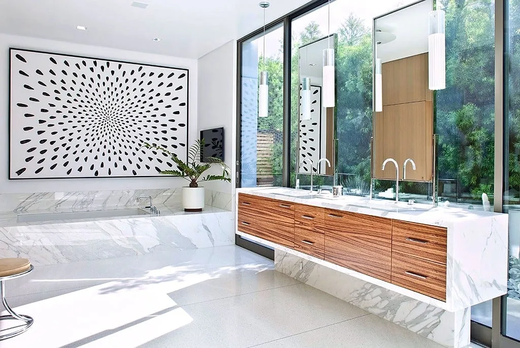 Zebrawood floating double vanity with a mitred white marble waterfall top runs the length of a glass-walled marble bathroom, facing a sunken marble tub deck and full-height windows