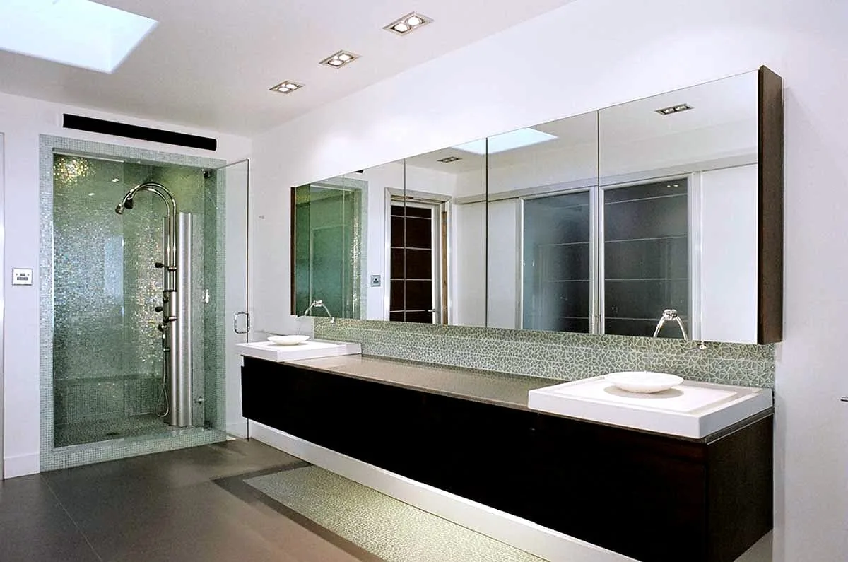 Long espresso floating vanity with twin square vessel basins and a glass mosaic backsplash runs beneath a full-width mirrored cabinet wall, beside a glass shower under a skylight