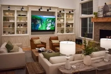 Floor-to-ceiling cream built-in media wall with X-mullion lower doors, open display shelving and integrated picture lights framing a wall-mounted television beside a brick fireplace