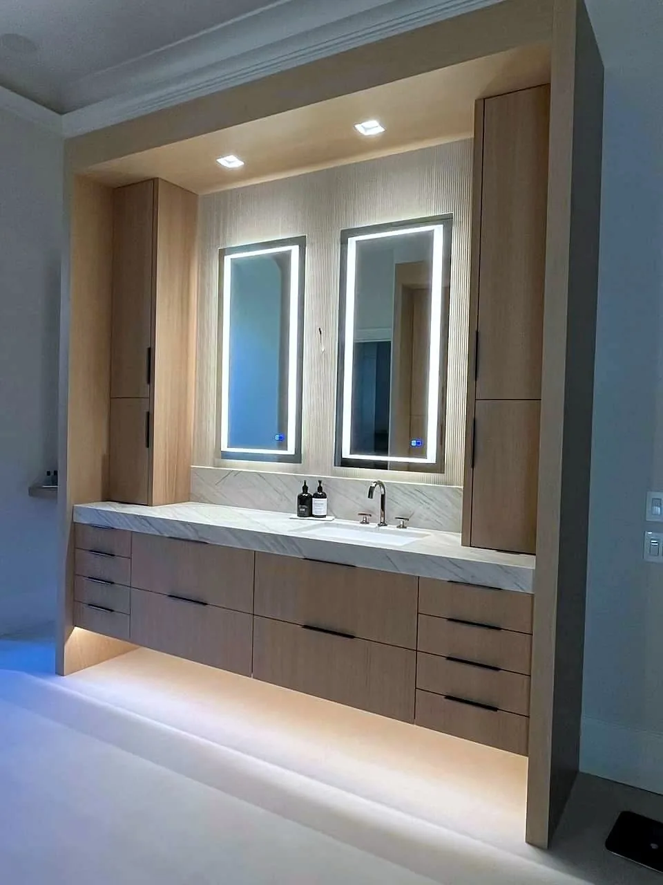 Full-wall floating vanity in light white oak veneer with horizontal-grain slab drawer banks and vertical-grain flanking towers, slim black bar pulls, veined white stone top with thick mitred edge and slab splash, single undermount rectangular sink, backlit LED mirrors and under-cabinet LED