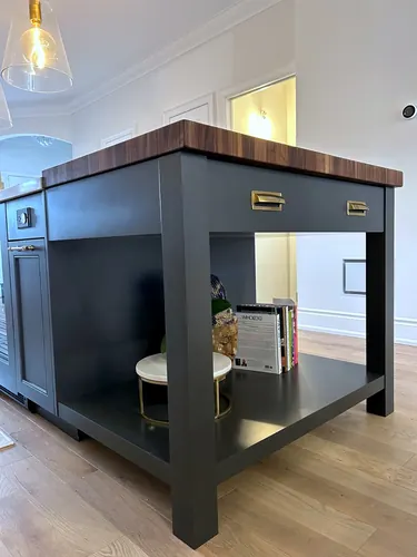 A dark charcoal island topped with a chunky walnut butcher-block counter and brass cup-pull drawers, an open lower display shelf, and a raised-panel end cabinet on light oak flooring — custom kitchen hand-built by Classic Woodworking & Vanity in Oakville, Ontario