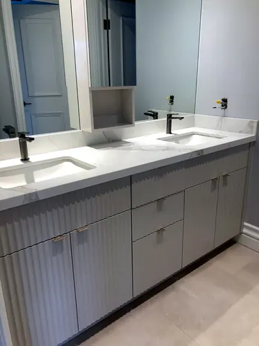 White fluted double vanity with a Calacatta marble-look waterfall counter, dual undermount sinks, brushed-nickel pulls, black faucets and an open cubby niche — custom vanity hand-built by Classic Woodworking & Vanity in Toronto, Ontario