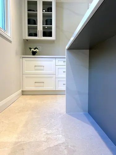 A bright pantry corner with white shaker drawers and glass-front upper cabinet holding dark dishware meets a waterfall marble-look quartz counter, brushed nickel bar pulls and a vase of daisies — custom kitchen hand-built by Classic Woodworking & Vanity in Markham, Ontario