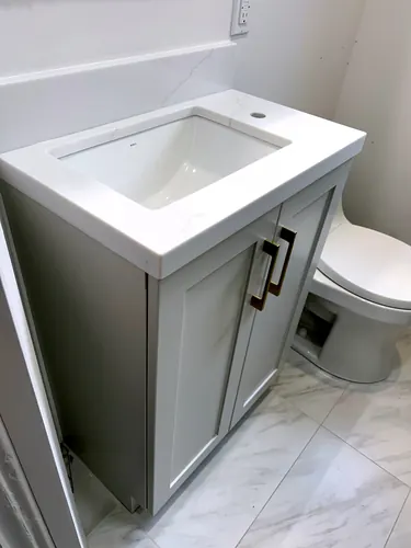 A compact light grey shaker vanity with brushed gold square bar pulls sits under a white quartz counter with integrated basin and matching backsplash, beside a white toilet on marble-look tile — custom vanity hand-built by Classic Woodworking & Vanity in Guelph, Ontario