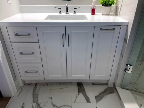 A white raised-panel vanity with polished chrome bar pulls carries a white quartz counter with undermount sink and chrome widespread faucet, set on dramatic white-and-grey marble tile beside a glass shower — custom vanity hand-built by Classic Woodworking & Vanity in Cambridge, Ontario