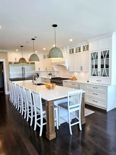 Long white shaker kitchen with X-mullion glass upper cabinets, a sculpted plaster range hood and marble-look quartz counters, anchored by an entertaining island with timber post legs and oversized dome pendants