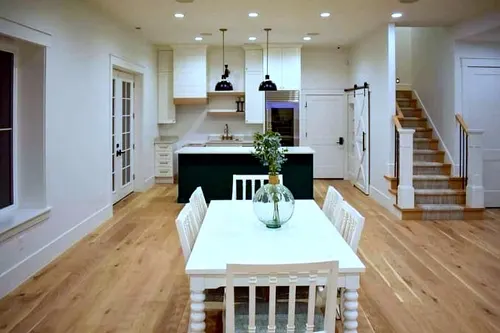 Open-plan white kitchen with a black-painted island, brass gooseneck faucet and black dome pendants, opening to a dining area with barn-door pantry and light oak flooring throughout