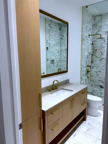 A light oak vanity with slab drawers, brass bar pulls and an open towel shelf beneath a white terrazzo-look counter, brass gooseneck faucet and brass-framed mirror beside a marble-tiled walk-in shower — custom vanity hand-built by Classic Woodworking & Vanity in Burlington, Ontario