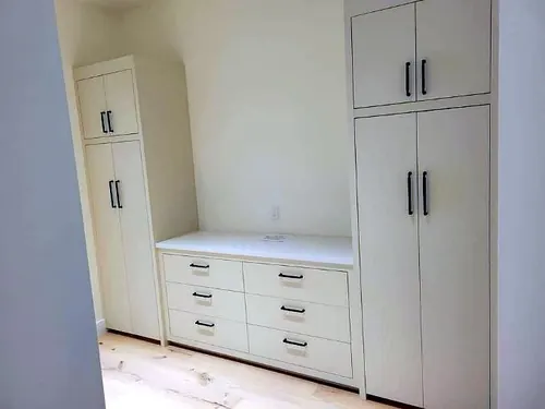 A run of painted cream Shaker cabinetry with tall pantry towers flanking a lower six-drawer bank and a white counter, all fitted with matte black bar pulls and knobs over pale wood flooring