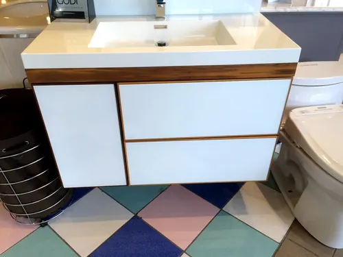 A showroom floating vanity with glossy white glass drawer fronts, natural wood edge banding and a thick white integrated basin top displayed above a diamond-tiled floor