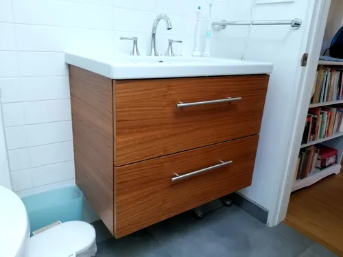 A warm walnut floating vanity with two drawers and long stainless bar pulls, a white drop-in sink and widespread chrome faucet beside a white-tiled wall