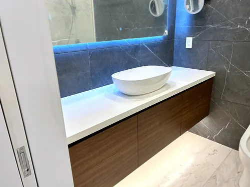 Floating walnut vanity with flat-slab doors and drawers, white quartz top holding a white oval vessel basin, backed by grey marble tile and a backlit LED mirror