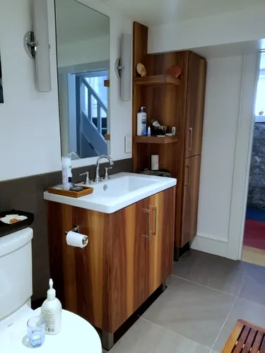 Curved-front walnut vanity with chrome bar pull and a wide white drop-in sink paired with a full-height walnut linen tower and open display shelves, chrome faucet and sconce over grey wainscot tile — custom vanity hand-built by Classic Woodworking & Vanity in Vaughan, Ontario
