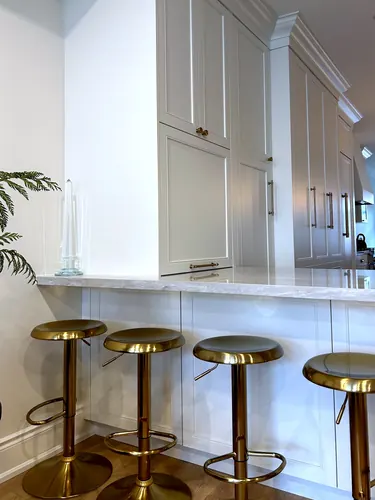 Floor-to-ceiling greige inset-panel kitchen cabinetry with brushed gold bar pulls and knobs meeting a light marble waterfall breakfast bar lined with four brass swivel stools over oak flooring