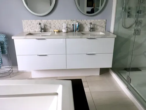 Wall-mounted white slab four-drawer vanity with slim chrome edge pulls, marble-look waterfall quartz top and twin oval mirrors above a textured stone mosaic backsplash beside a glass shower