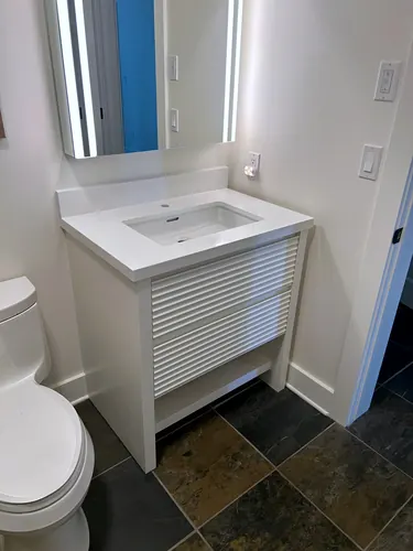 Freestanding white vanity with reeded fluted drawer fronts, an integrated white quartz sink-top and backlit mirror, set against multicoloured slate floor tile beside the toilet — custom vanity hand-built by Classic Woodworking & Vanity in Guelph, Ontario