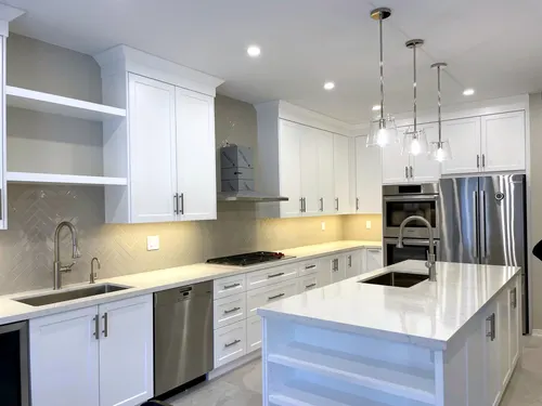 Bright white shaker kitchen with satin nickel bar pulls, herringbone tile backsplash and white quartz counters, a stacked stainless wall oven and integrated dishwasher, and glass pendants over the island prep sink