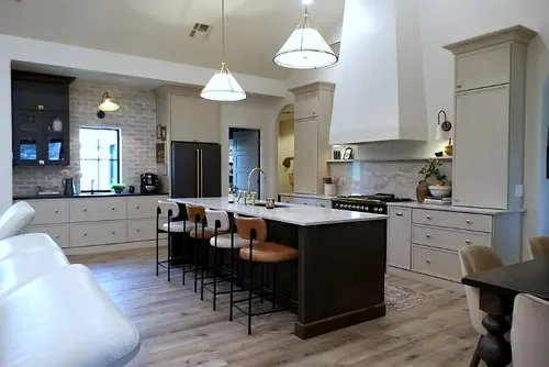 Cream perimeter cabinetry with a plaster range hood and brick-look backsplash pairs with a dark stained island and marble-look quartz top, seated with leather counter stools under linen drum pendants