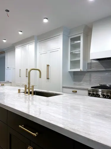 Creamy white inset cabinetry with brass bar pulls, a reeded-glass upper cabinet, marble backsplash and range hood, a brass bridge faucet at the undermount sink, and a dark island with quartzite counter — custom kitchen hand-built by Classic Woodworking & Vanity in Etobicoke, Ontario