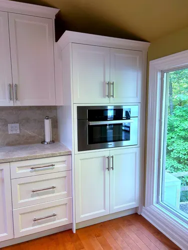 White inset cabinetry with a built-in stainless Miele wall oven, satin nickel bar pulls, a stone slab backsplash and counter, and drawer bank beside a bright garden window over maple flooring