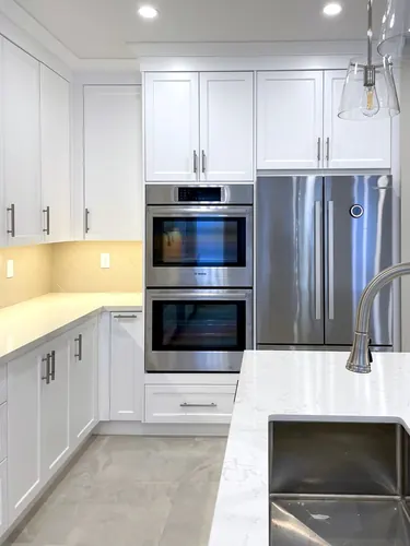 Bright white shaker kitchen with a stacked stainless Bosch double wall oven, panel-fronted French-door fridge, herringbone tile backsplash, satin nickel pulls, and marble-look quartz counters on grey tile — custom kitchen hand-built by Classic Woodworking & Vanity in Toronto, Ontario